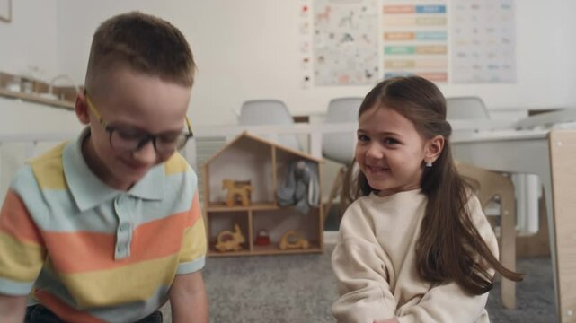 Close-up tilt of young Caucasian girl and boy sitting on carpet floor at nursery playing numeracy card game and giving high-five to each other after completing task