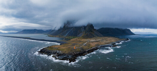 Stunning aerial view of Vestrahorn mountaine on Stokksnes cape in Iceland's rugged coastline featuring a small orange lighthouse on a rocky peninsula, black sand beach