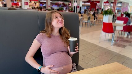 Young happy pregnant woman drinking coffee on food court in shopping mall. Pregnancy and caffeine. 