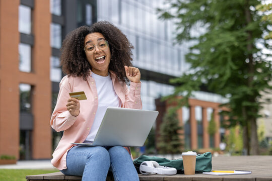 Happy young female student holding credit card while using laptop outdoors near university campus. Concept of online shopping, education, and student life.
