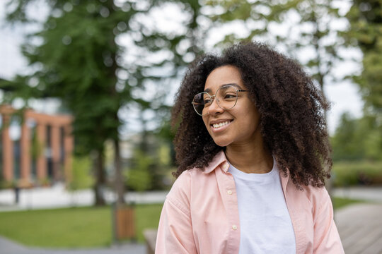 Young female student smiling while standing outdoors near university campus. Natural and relaxed setting with greenery and buildings in the background. Ideal for themes of education and campus life.