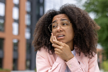 Woman with curly hair and glasses experiencing toothache outdoors. She looks uncomfortable and in...