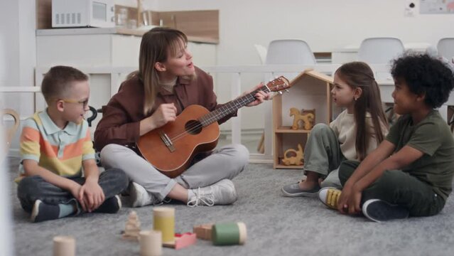 Full shot of Caucasian female educator sitting on floor in classroom playing guitar and singing song in company of diverse smiling preschool children trying to keep rhythm