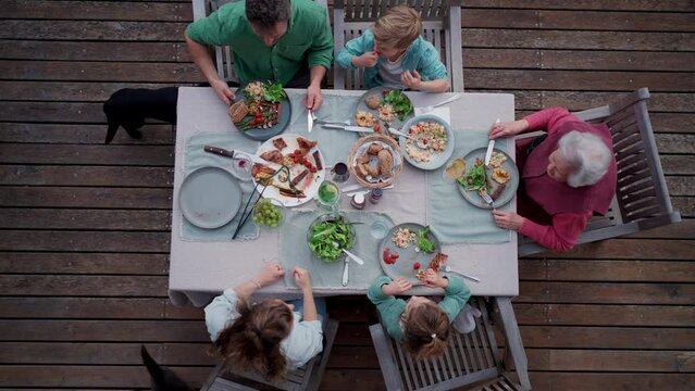 Top view of multigenerational family sitting at big dining table, enjoying grilled food. Family barbecue in backyard, spending quality time outdoors together.