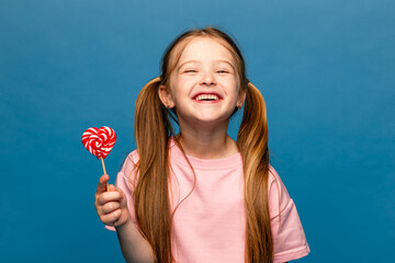 Happiness, childhood, kid and sweets. A happy little girl holding a heart shaped lollipop caramel on a stick, A festive candy in the hands of child in pink t-shirt on blue background. Valentines Day
