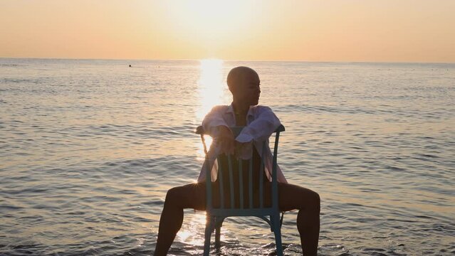 beautiful women with alopecia on beach against sea drag a chair.balt girl in shirt in water, sunrise or sunset time.sun reflection on waves.female in swimsuit.