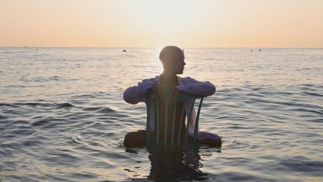 beautiful women with alopecia on beach against sea drag a chair.balt girl in shirt in water, sunrise or sunset time.sun reflection on waves.female in swimsuit.