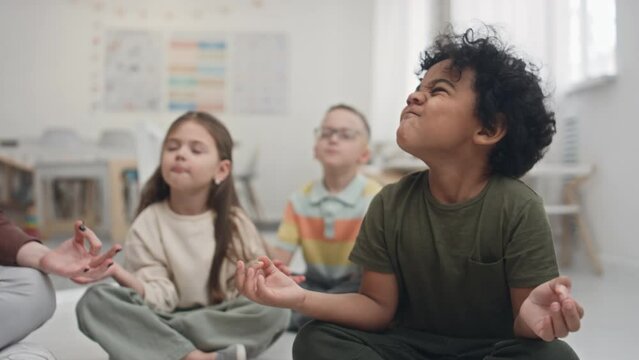 Medium shot of young multiethnic children sitting on floor trying to stay calm during breathing and self-regulating practice. Offscreen female educator helping kids to relax