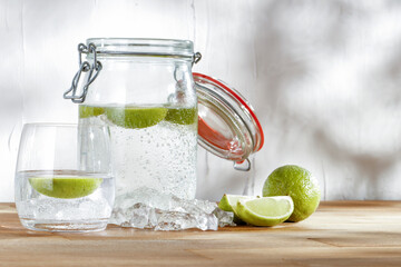 Water with lime in a glass jar and a glass on a wooden table against a white wall and natural light. Advertising space