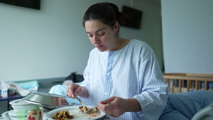 Woman in a hospital gown eating a meal on a tray while sitting on a hospital bed, showing the care and support provided to patients during their recovery period in the hospital