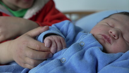 Young sibling gently holding the hand of a sleeping newborn baby, showcasing family love, early...