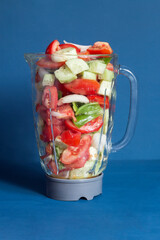A clear glass blender filled with fresh, sliced vegetables including tomatoes, cucumbers, and bell peppers against a vibrant blue background highlights healthy eating and food preparation.