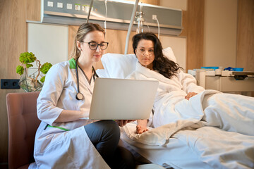 Specialist sits near a woman on hospital bed with laptop