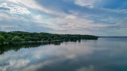 A blue sky with clouds and a shore with green trees reflected in the lake. Beautiful summer nature is mirrored in the water. Drone shot of the landscape, aerial view photo.