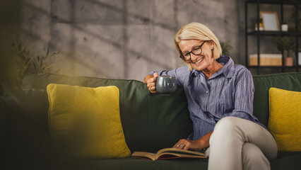 Mature woman sit in the living room read a book and drink coffee