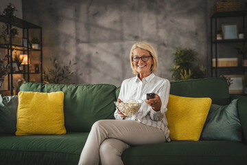Obraz premium Mature senior woman sit on sofa hold popcorn and watch movie at home
