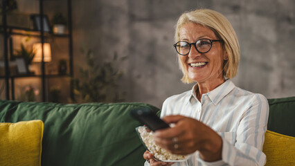 Mature senior woman sit on sofa hold popcorn and watch movie at home