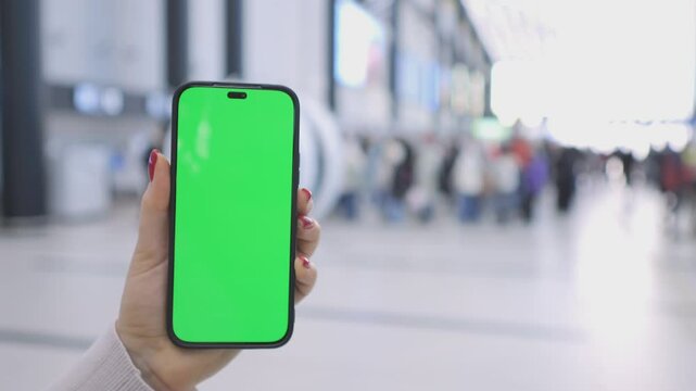 A person holding a smartphone with a green screen in a crowded airport terminal