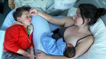 Obraz premium Mother breastfeeding newborn while comforting her older son in a red hoodie, highlighting the warmth and connection within a family, depicting love, care, and maternal devotion