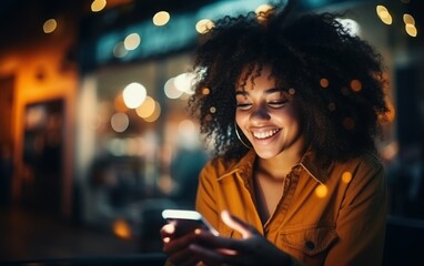 A young African American woman with curly hair smiles while using her smartphone in an urban setting at night