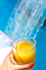 A glass of orange lemonade with a slice of orange and ice in hand against the background of blue pool water and water jets