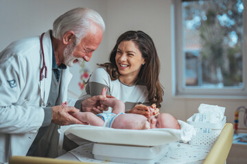 A smiling elderly male pediatrician in a white coat examines a baby with a joyful young adult female observing, in a well-equipped hospital room.