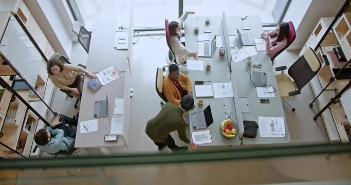 Overhead view of a group of people engaging in discussions and strategizing in a well-lit, open-plan office setting. Team members are seen at separate desks, collaborating and working on laptops.