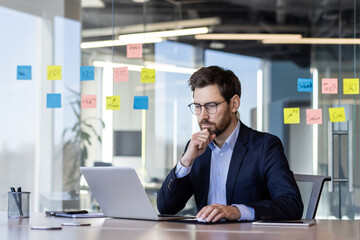 Businessman working on laptop in modern office with sticky notes on glass wall. Focused...