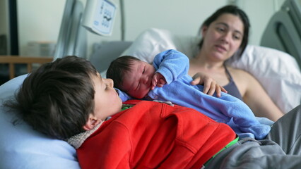 Newborn baby in blue onesie cuddling with older sibling, both resting on hospital bed, with mother watching lovingly in the background, capturing a warm and affectionate family moment