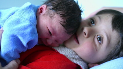 Newborn baby resting on older brother's chest, both snuggled closely, with baby alert and looking...