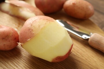 Fresh raw potatoes, peels and peeler on wooden table, closeup