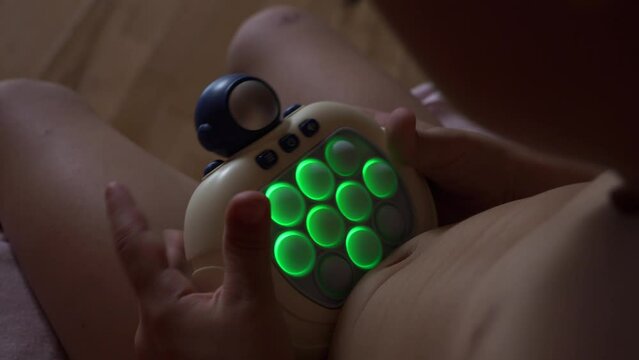 A little boy plays with an electronic pop light-up fidget toy at home, over the shoulder shot.