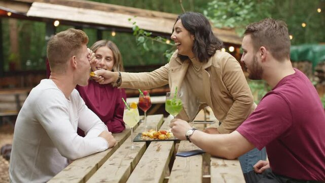 Four young friends enjoying outdoor meal with drinks, appetizers, in rustic setting - woman is playfully feeding man - everyone is smiling and laughing - lively and enjoyable moment
