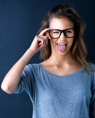 Wink portrait, tongue and teenager girl with glasses for vision, funny expression and feeling goofy in studio. Emoji face, gen z or young woman with cool eyewear, comedy and happy by dark background