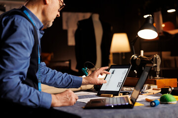 Elderly tailor picking perfect handdrawn outfit design on tablet to use as inspiration for customer comission. Experienced suitmaker creating custom made sartorial clothing in atelier