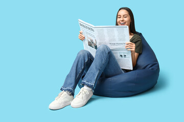 Beautiful young woman in beanbag chair with newspaper on blue background