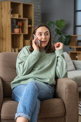 Obraz premium Happy young woman talking by mobile phone in armchair at home