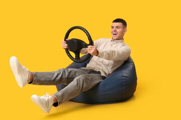 Happy young man with steering wheel sitting on beanbag chair against yellow background