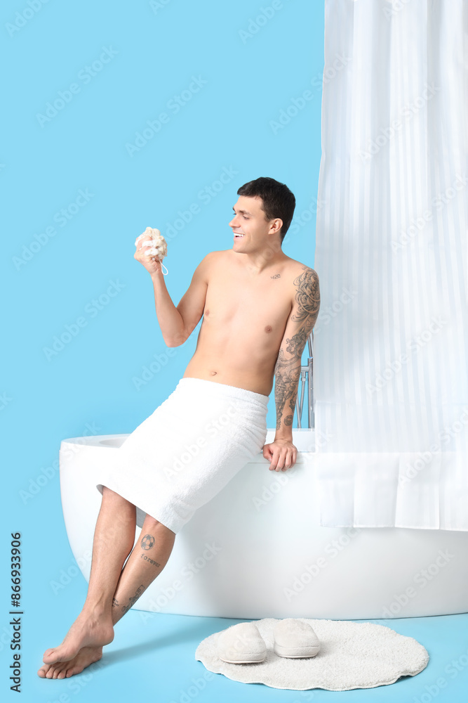 Wall mural Young man with loofah sitting on bathtub against blue background