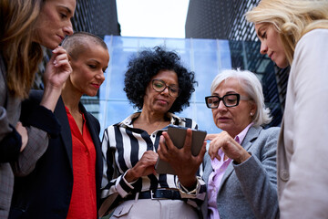 Low angle. Team of diverse ages and races business women gathered standing concentrated using and holding tablet pc outdoor. Entrepreneur females serious looking screen device outside work building