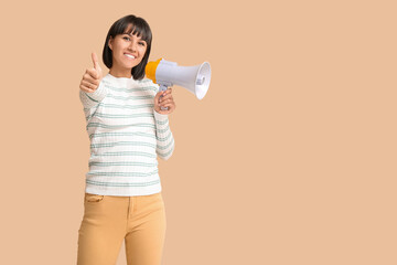 Young woman with megaphone showing thumb-up on  beige background