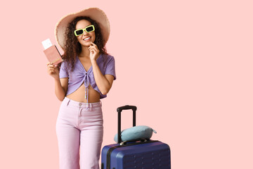 Happy young African-American woman with suitcase, passport and travel pillow on pink background