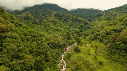 Fototapeta premium AERIAL Hazy hilltops above beautiful lush rainforest with flowing mountain river