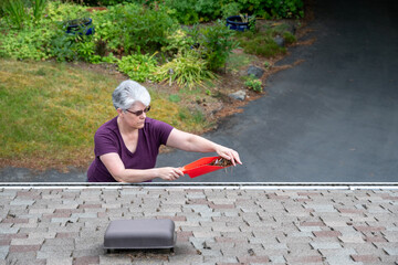 Middle aged white woman with gray hair cleaning out pine needles and leaf debris from a roof rain...