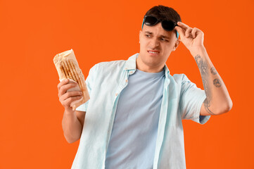 Handsome young man with skeptical expression holding tasty doner kebab on orange background