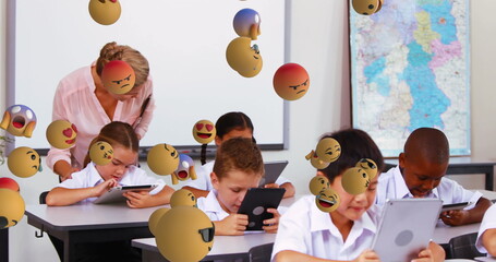 Image of emoticons floating over diverse female teacher helping students with digital tablet