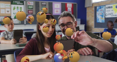 Image of emoticons over diverse male teacher helping girl in using digital tablet in classroom