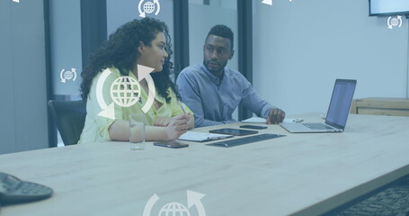 Image of globe icons in arrows over diverse coworkers discussing reports on laptop in office