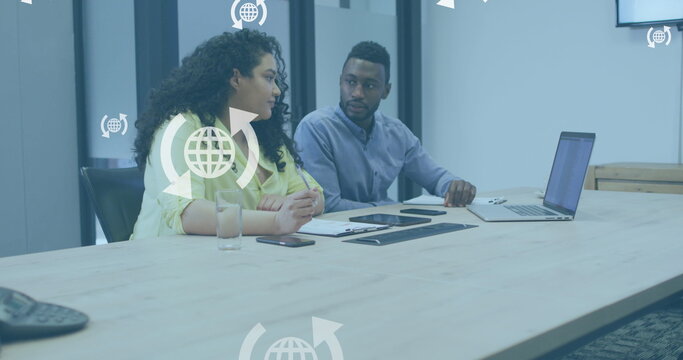 Image of globe icons in arrows over diverse coworkers discussing reports on laptop in office