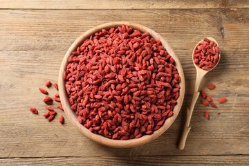 Dried goji berries in bowl and spoon on wooden table, flat lay